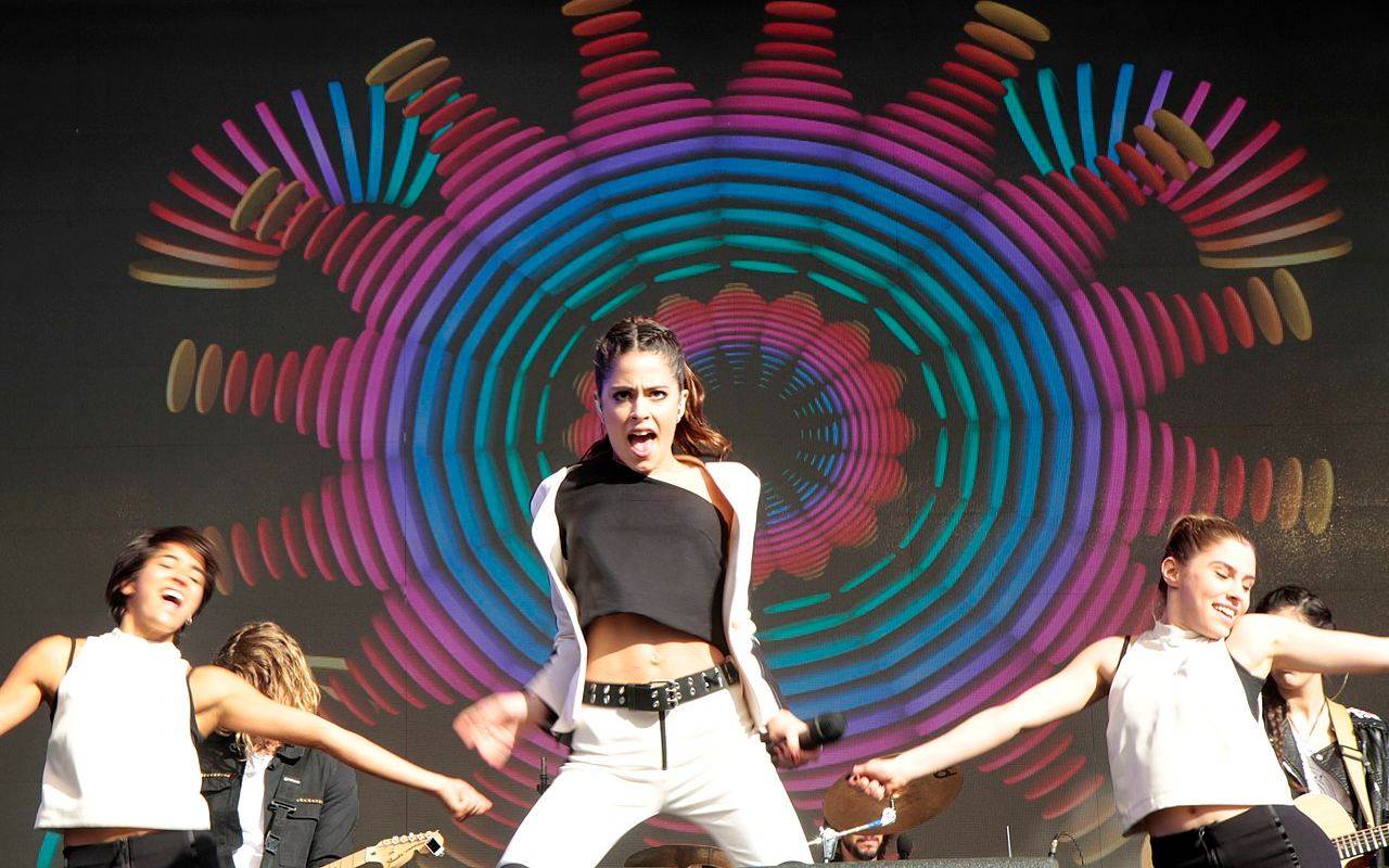 Tini Stoessel cantando y bailando en un escenario con bailarines de fondo y una pantalla colorida de fondo