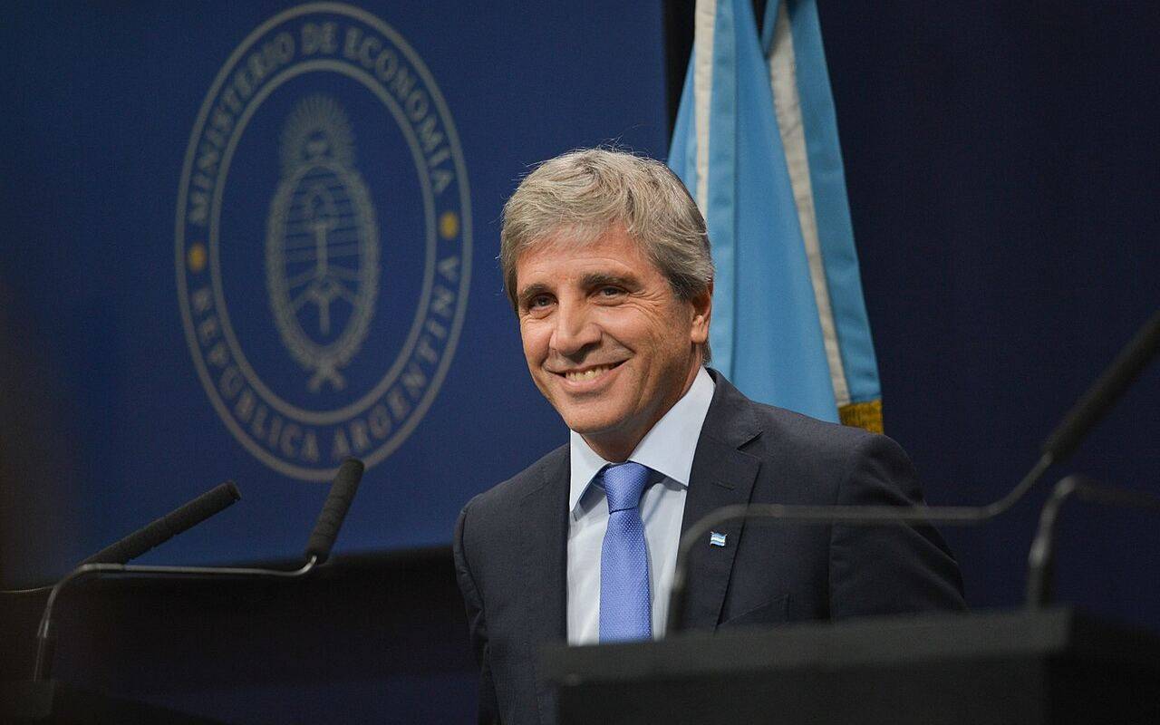 Luis Caputo, de traje y corbata, detrás de un atril con micrófonos y la bandera argentina al fondo.